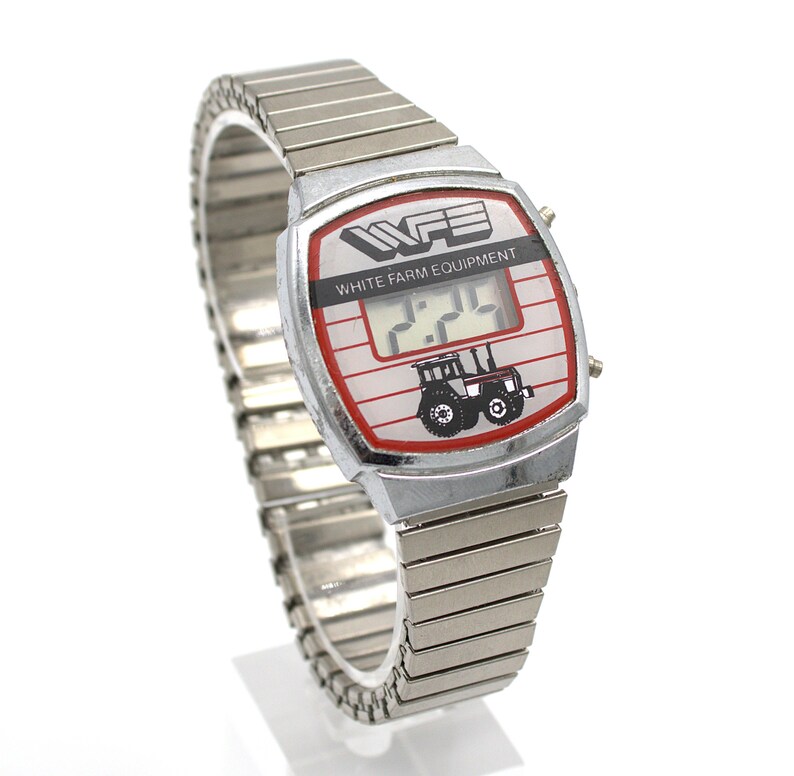 Peut inclure: Montre num&eacute;rique argent&eacute;e avec un cadran rectangulaire. Le cadran affiche le texte "WFE WHITE FARM EQUIPMENT" et un dessin de tracteur. L'heure affich&eacute;e est 2:24. Le bracelet est en m&eacute;tal argent&eacute; flexible. La montre mesure environ 3 cm de large.