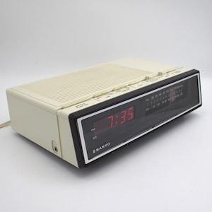 May include: A vintage Sanyo digital alarm clock radio. The clock displays 7:35 in red digits. The radio has a cream-colored body with a black and silver face. The Sanyo logo is visible.