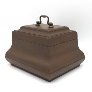 May include: A brown wooden box with a hinged lid and a metal handle. The box has a slightly curved shape and a smooth finish.