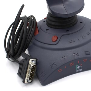 Puede incluir: Un joystick Logitech Extreme Digital gris oscuro con cable y conector negros. El joystick tiene botones rojos y el texto "Extreme Digital" y "Wingman" impresos.