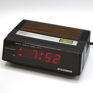 May include: A black digital alarm clock with a red LED display showing the time 7:52 PM. The clock has a silver face with the text "DISPLAY MADE IN TAIWAN R.O.C." and "AUTO ALARM PM". The clock is made by SYLVANIA.