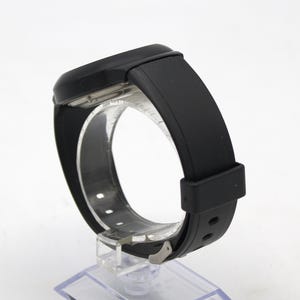 May include: A black wristwatch with a rectangular face and a black rubber strap. The watch is displayed on a clear plastic stand, showcasing the band's design and the watch's overall sleek appearance.