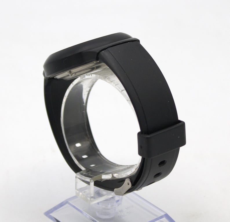May include: A black wristwatch with a rectangular face and a black rubber strap. The watch is displayed on a clear plastic stand, showcasing the band's design and the watch's overall sleek appearance.