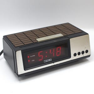 May include: A black digital alarm clock with a red LED display showing the time 5:48. The clock has a brown woodgrain top and a silver trim. The clock has a COSMO brand name.