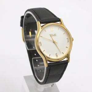 May include: A gold-toned wristwatch with a white face and black leather band. The watch face displays the word "TIME" and the numbers 3, 6, 9, and 12. The watch is displayed on a clear stand.