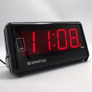 May include: Black digital alarm clock displaying 11:08 PM in bright red digits. The clock has a rectangular shape with a black frame and the brand name "Spartus" printed on the front. The alarm clock is on a white surface.