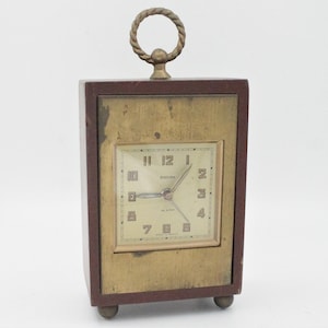 May include: A vintage wooden alarm clock with a brass top and a gold-colored face. The clock face has black Roman numeral hour markers and black hands. The clock face also has the word "Engoura" printed on it.