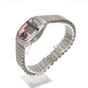 Peut inclure: Montre num&eacute;rique argent&eacute;e avec un cadran rectangulaire. Le cadran pr&eacute;sente un design rouge et blanc avec le texte "WHITE FARM EQUIPMENT" et un dessin de tracteur. La montre a un bracelet en m&eacute;tal argent&eacute;.