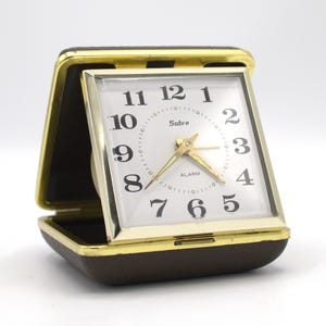 May include: A vintage Sabre travel alarm clock with a gold-tone frame and a brown leatherette case. The clock face is white with black numerals and gold hands. The clock is open, revealing the face and the alarm setting.