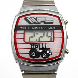 Peut inclure: Montre num&eacute;rique vintage avec bracelet et bo&icirc;tier en m&eacute;tal argent&eacute;. Le cadran affiche "WHITE FARM EQUIPMENT" au-dessus d'un affichage num&eacute;rique de l'heure. Une bordure rouge entoure le cadran, avec un dessin de tracteur en bas.
