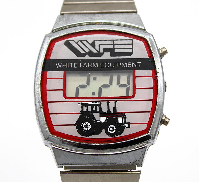 Peut inclure: Montre num&eacute;rique vintage avec bracelet et bo&icirc;tier en m&eacute;tal argent&eacute;. Le cadran affiche "WHITE FARM EQUIPMENT" au-dessus d'un affichage num&eacute;rique de l'heure. Une bordure rouge entoure le cadran, avec un dessin de tracteur en bas.