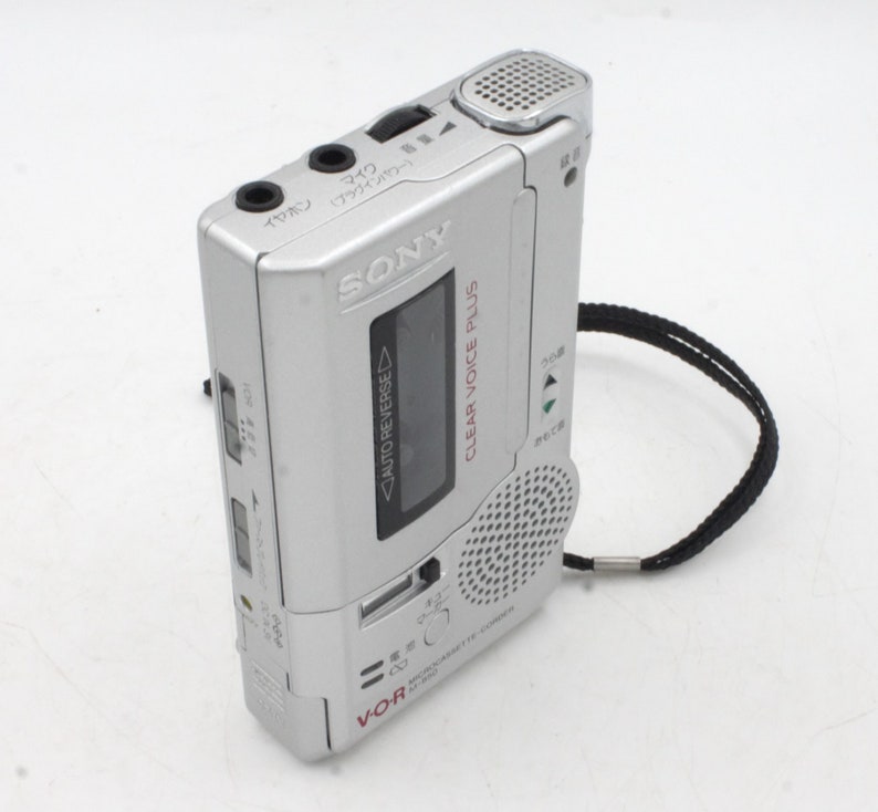 May include: A silver Sony Clear Voice Plus voice recorder with a black strap. The recorder has a small LCD screen, a microphone, and a speaker. The text "VOR" and "AUTO REVERSE" are visible on the recorder.