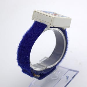 May include: A vintage children's watch with a white rectangular face featuring a colorful design. The watch has a blue, fuzzy Velcro strap. The watch is displayed on a clear plastic stand.
