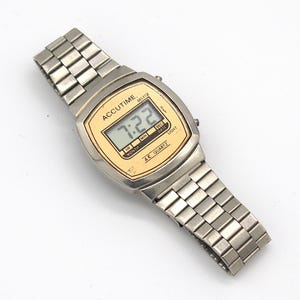 May include: A silver Accutime digital watch with a gold-toned face displaying the time 7:22. The watch has a rectangular face and a silver link band. The watch face has the words "ACCUTIME" and "QUARTZ".