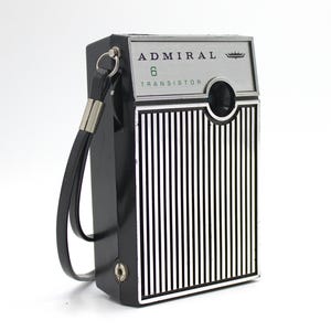 May include: A black and silver vintage Admiral 6 transistor radio with a carrying strap. The radio has a silver grill with vertical lines and a round speaker.