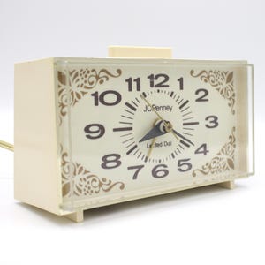 May include: A white rectangular alarm clock with a gold-colored face and black numbers. The clock has a decorative floral design around the edge of the face. The clock face says "JCPenney Lighted Dial".