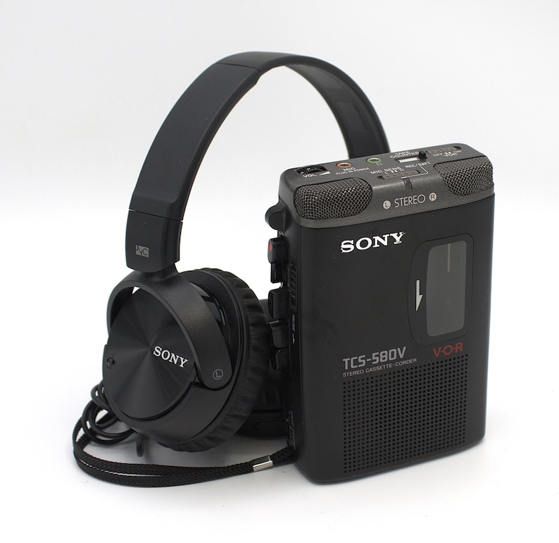 May include: Black Sony headphones and a black Sony TCS-580V stereo cassette-corder. The cassette recorder has the text "STEREO" and "V-O-R" printed on it. The headphones are connected to the recorder with a black cord.