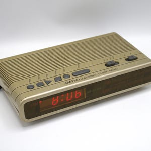 May include: A gold-colored AM/FM electronic clock radio displaying 8:06 in red digital numbers. The rectangular device has several buttons and a ridged top. The text "AM FM ELECTRONIC CLOCK RADIO" is printed on the front.