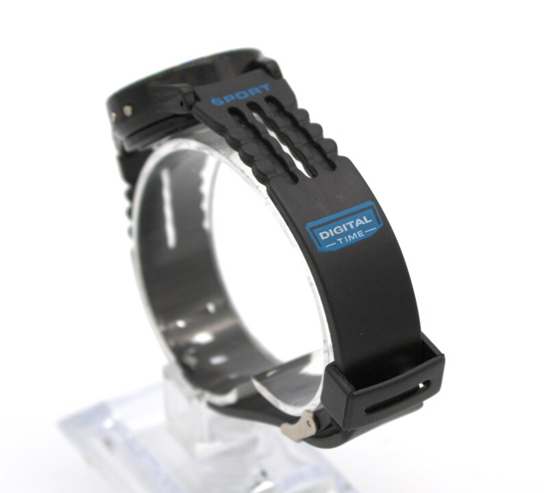 May include: Black digital sport watch with a black band. The watch face is not visible. The band has the word "SPORT" printed in blue and the words "DIGITAL - TIME" printed in blue.