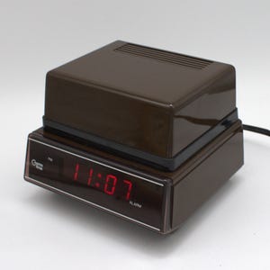 May include: A brown Cosmo Time digital alarm clock with a dark, translucent front display panel. The clock features a bright red LED display showing the time 11:07, with the text Cosmo Time and ALARM printed in white on the front face.