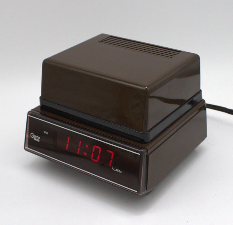 May include: A brown Cosmo Time digital alarm clock with a dark, translucent front display panel. The clock features a bright red LED display showing the time 11:07, with the text Cosmo Time and ALARM printed in white on the front face.