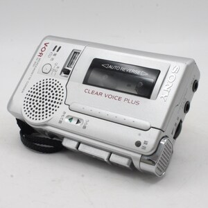 May include: A silver Sony Microcassette recorder with a black strap. The recorder has a built-in speaker, a clear voice plus feature, and auto reverse functionality. The text "V-O-R Microcassette-Corder 14-850" is printed on the side.