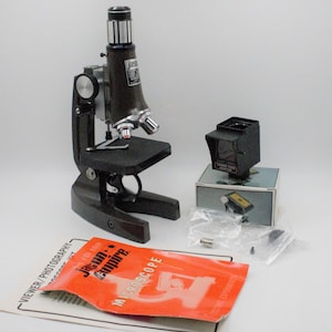 May include: A black and silver microscope with a viewer/photography accessory. The microscope has a black base and a silver body with a black lens. The accessory is a black box with a red viewer and the text "VIEWER-TUBE ADAPTER". The microscope is on a white surface with a red and white instruction booklet.
