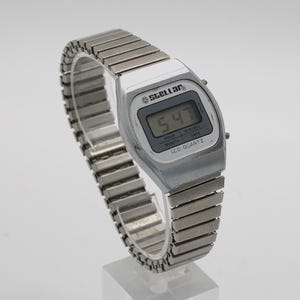 May include: A silver-toned digital wristwatch with a metallic expansion band. The watch face features the brand name Stellar, a digital display showing 5:47, and the text LCD QUARTZ, along with labels for HOUR, MONTH, MIN/SEC, and DATE.