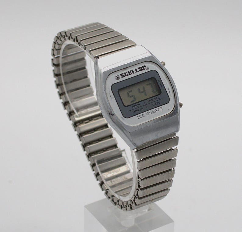 May include: A silver-toned digital wristwatch with a metallic expansion band. The watch face features the brand name Stellar, a digital display showing 5:47, and the text LCD QUARTZ, along with labels for HOUR, MONTH, MIN/SEC, and DATE.
