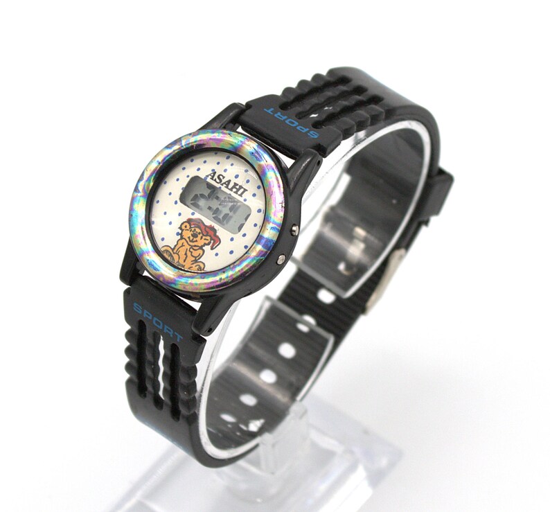 May include: Black ASAHI Sport watch with a digital display. The watch face has a colorful iridescent bezel and a cartoon bear graphic. The watch band is black with the word "SPORT" in blue.