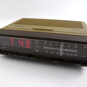 May include: A vintage brown and tan digital alarm clock displaying "7:49" in red. The clock features an FM/AM radio dial, volume and tuning controls, and a digital display. The clock is rectangular and has a retro design.