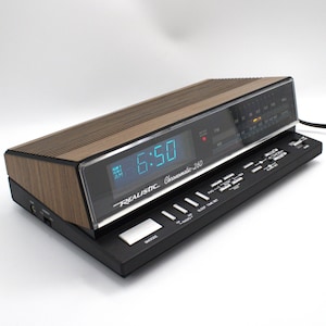 May include: A vintage Realistic Chronomatic-260 digital clock radio. The clock features a wood-grain top and a black base with control buttons. The digital display shows the time in blue numerals. The radio dial is visible behind a clear plastic cover.