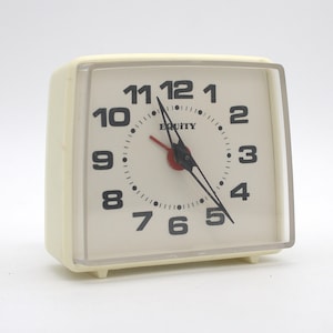 May include: A square-shaped off-white analog alarm clock with a clear plastic front cover. The clock face features black Arabic numerals from 1 to 12, black hour and minute hands, a red second hand, and the brand name Equity printed in black.