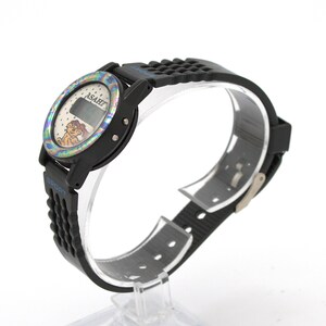 May include: Black digital watch with a colorful bezel and a black rubber strap. The watch face displays the brand "ASAHI" and a cartoon character. The strap has the word "SPORT" in blue.