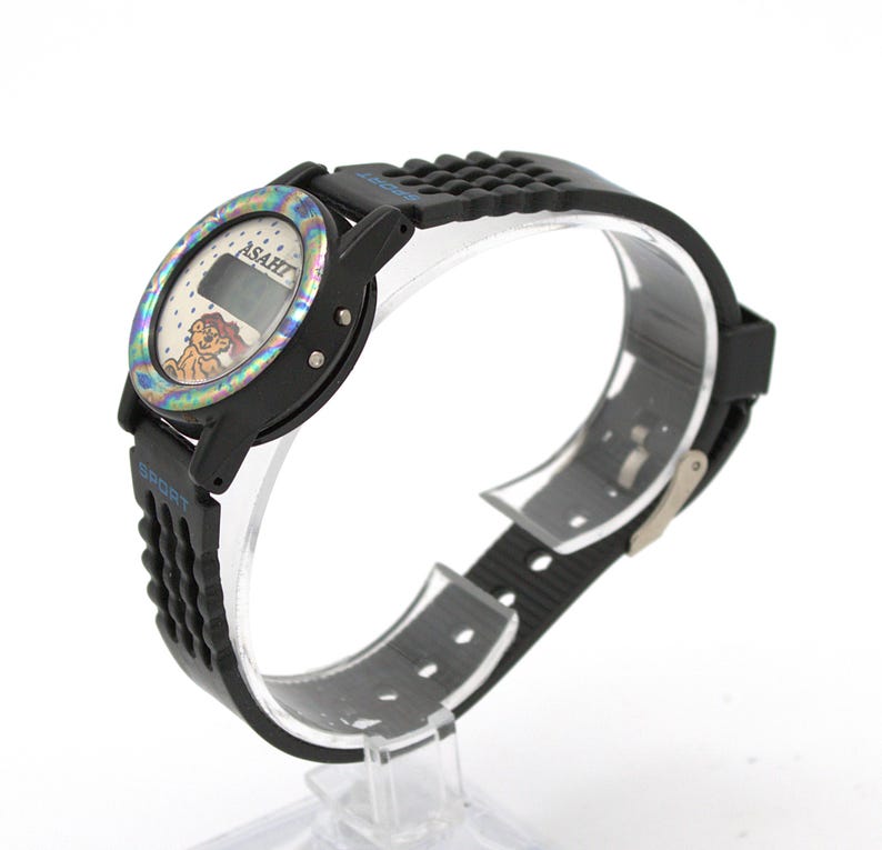 May include: Black digital watch with a colorful bezel and a black rubber strap. The watch face displays the brand "ASAHI" and a cartoon character. The strap has the word "SPORT" in blue.