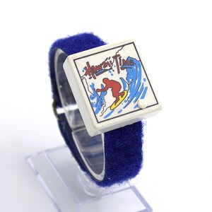 May include: A vintage watch with a blue Velcro strap and a white face. The face features the words "Hawaii Time" above a graphic of a surfer riding a wave. The watch is displayed on a clear plastic stand.
