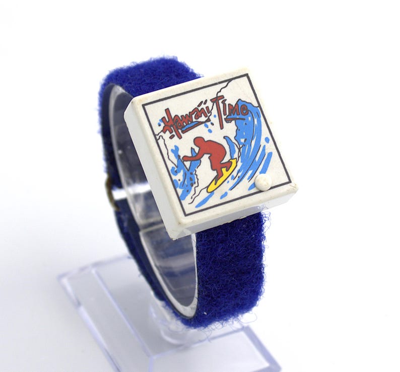 May include: A vintage watch with a blue Velcro strap and a white face. The face features the words "Hawaii Time" above a graphic of a surfer riding a wave. The watch is displayed on a clear plastic stand.