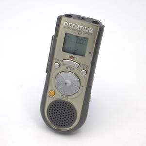 May include: A silver Olympus VN-1800 digital voice recorder with a black casing. The recorder has a small LCD screen, a circular control knob, and buttons for play, stop, record, menu, and folder.