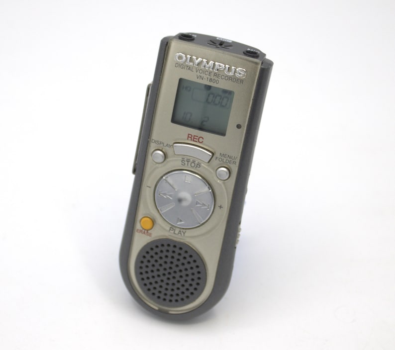May include: A silver Olympus VN-1800 digital voice recorder with a black casing. The recorder has a small LCD screen, a circular control knob, and buttons for play, stop, record, menu, and folder.