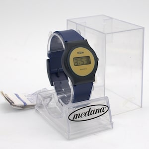 May include: A navy blue digital wristwatch with a round black bezel and a gold-toned face. The watch features the text Medana and Quartz on the display. It is presented on a clear plastic display stand with the Medana logo and includes a clear box.