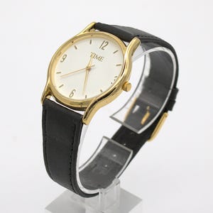 May include: A gold-toned wristwatch with a white face and black leather band. The watch face displays the word "TIME" and the numbers 12, 6, and 9. The watch is displayed on a clear stand.