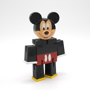May include: A block-style Mickey Mouse figure constructed from geometric shapes. The character has a black head with round ears, a tan face, and a black torso. It wears red shorts with two white oval buttons, black legs, and yellow shoes.
