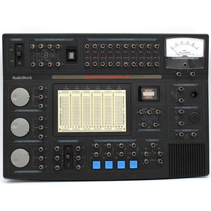 May include: A black RadioShack Electronics Learning Lab featuring a central breadboard, three gray potentiometers, four blue push-button switches, a DC milliammeter, various LED indicators, and multiple input and output connection ports.