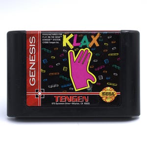 May include: A black Sega Genesis video game cartridge for the game Klax. The label features a pink hand graphic, the game title in colorful letters, and the Sega logo. The cartridge is labeled "Tengen."