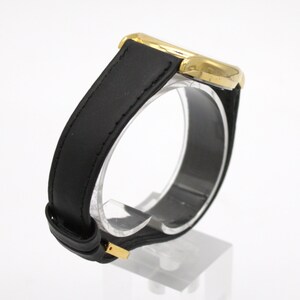 May include: A gold-toned wristwatch with a black leather band. The watch has a rectangular face and a classic design. The band is secured with a gold-colored buckle. The watch is displayed on a clear stand.