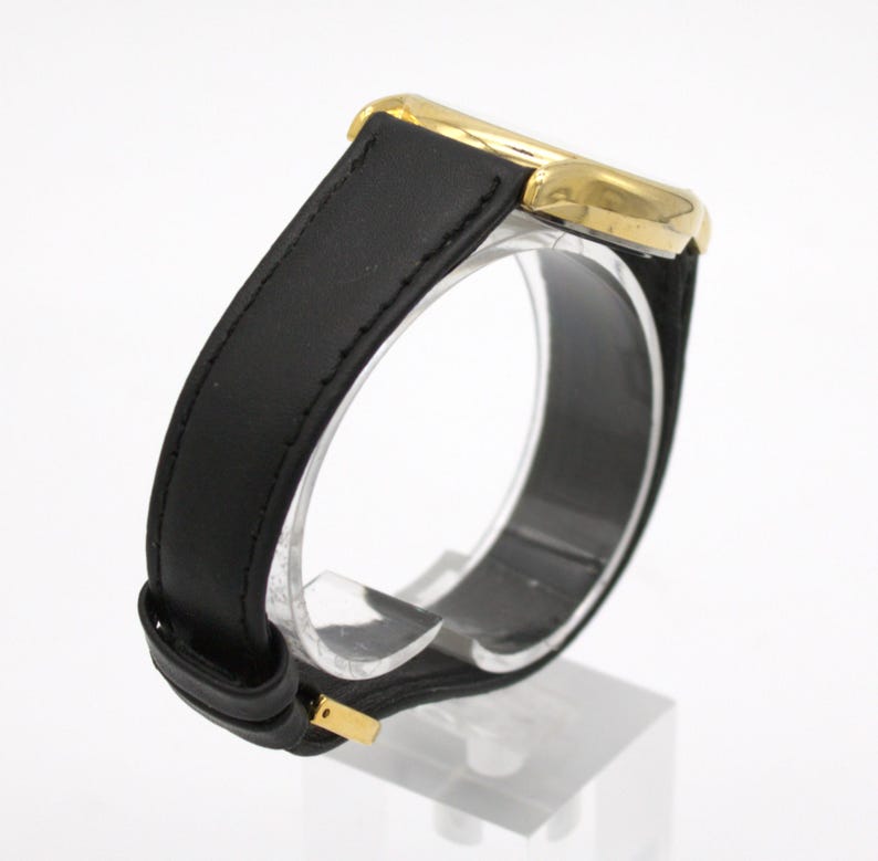 May include: A gold-toned wristwatch with a black leather band. The watch has a rectangular face and a classic design. The band is secured with a gold-colored buckle. The watch is displayed on a clear stand.
