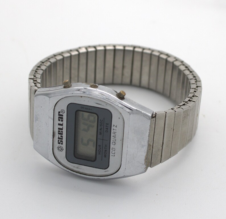 May include: A silver-tone digital wristwatch with an expandable metal link band. The watch face features a rectangular LCD screen displaying the time, with the brand name Stellar and the text LCD QUARTZ, HOUR, MIN/SEC, DATE, and MONTH printed on it.