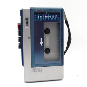 May include: A vintage Sony FM/AM cassette player in silver, blue, and gray. The player has a cassette compartment, a radio dial, and a black carrying strap. The text "SONY" and "FM/AM" are visible on the device.