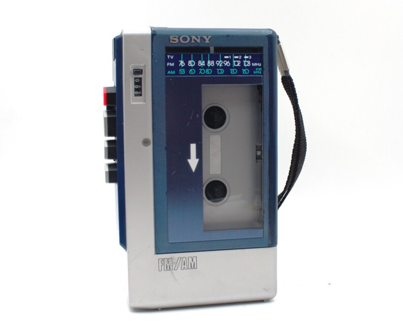 May include: A vintage Sony FM/AM cassette player in silver, blue, and gray. The player has a cassette compartment, a radio dial, and a black carrying strap. The text "SONY" and "FM/AM" are visible on the device.