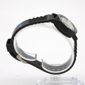 May include: Black wristwatch with a black band and a colorful face. The watch has a black bezel and a black band with a wave-like design. The watch is displayed on a clear stand.